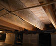 Wooden beams and wires under a house crawl space with concrete block supports, white mold patches, and a single light bulb.