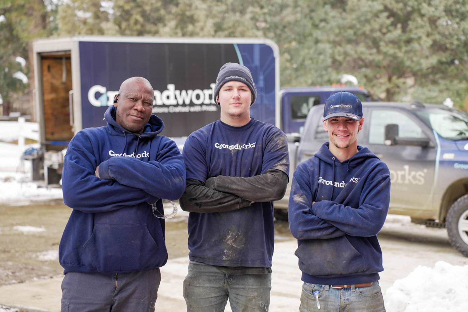 Three men in Groundworks uniforms stand with arms crossed in front of company trucks at a work site.