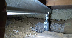 A steel support jack under a house in a crawl space with dirt, rocks, and foam insulation.
