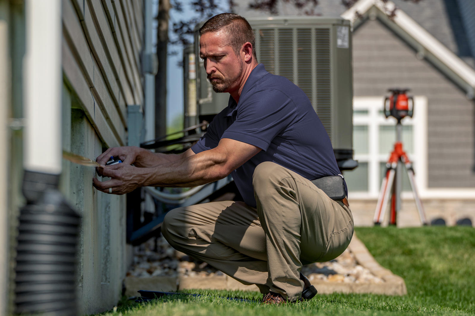 Man squatting outside a house, measuring the foundation wall with a tape measure.