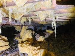 Yellow insulation hanging down and scattered in a dimly lit, cluttered crawl space under a house.