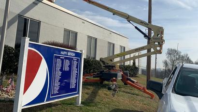 A lift is parked by a white building near a Pepsi birthday sign and a white truck on a grassy area.