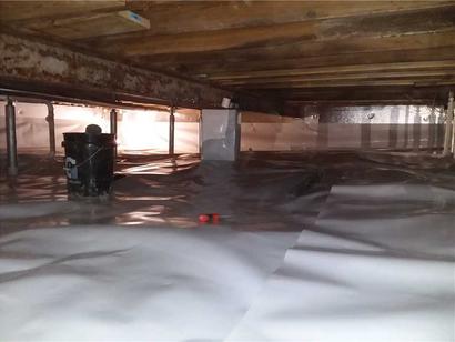 Crawl space with plastic vapor barrier on floor and supports; bucket and pipes are visible under wooden beams.