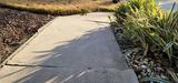 A concrete sidewalk bordered by green plants and gravel in sunlight.