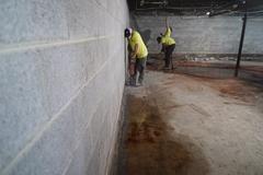 Two workers in yellow shirts use tools to break concrete along a basement wall with exposed pipes on the ceiling.