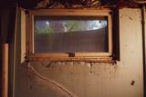 A small, dirty basement window with cracked, peeling walls and plants visible outside.