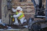 A construction worker in safety gear drills into the ground near a brick building with equipment nearby.