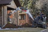 A worker operates a mini excavator, digging near a brick house while another worker observes.