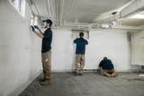 Three workers in uniforms install a white wall liner in a basement with exposed pipes and unfinished flooring.