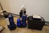 Two blue sump pumps with pipes and a black battery backup system on a concrete basement floor.
