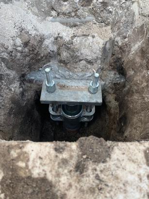 A close-up of a metal foundation pier installed in a dirt hole, secured with bolts and nuts.