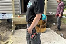 Man in work attire with tools stands outside a building under construction. Another person is in the background.