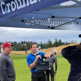 A man in a blue shirt interviews a golfer under a tent on a golf course, with others in the background.