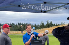 A man in a blue shirt interviews a golfer under a tent on a golf course, with others in the background.