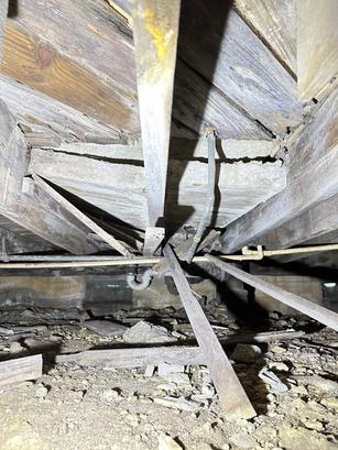 Damaged wooden floor joists and debris in a crawl space with exposed pipes and dirt ground.