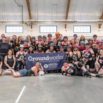A large roller derby team poses indoors, smiling together behind a "Groundworks" sponsor banner.
