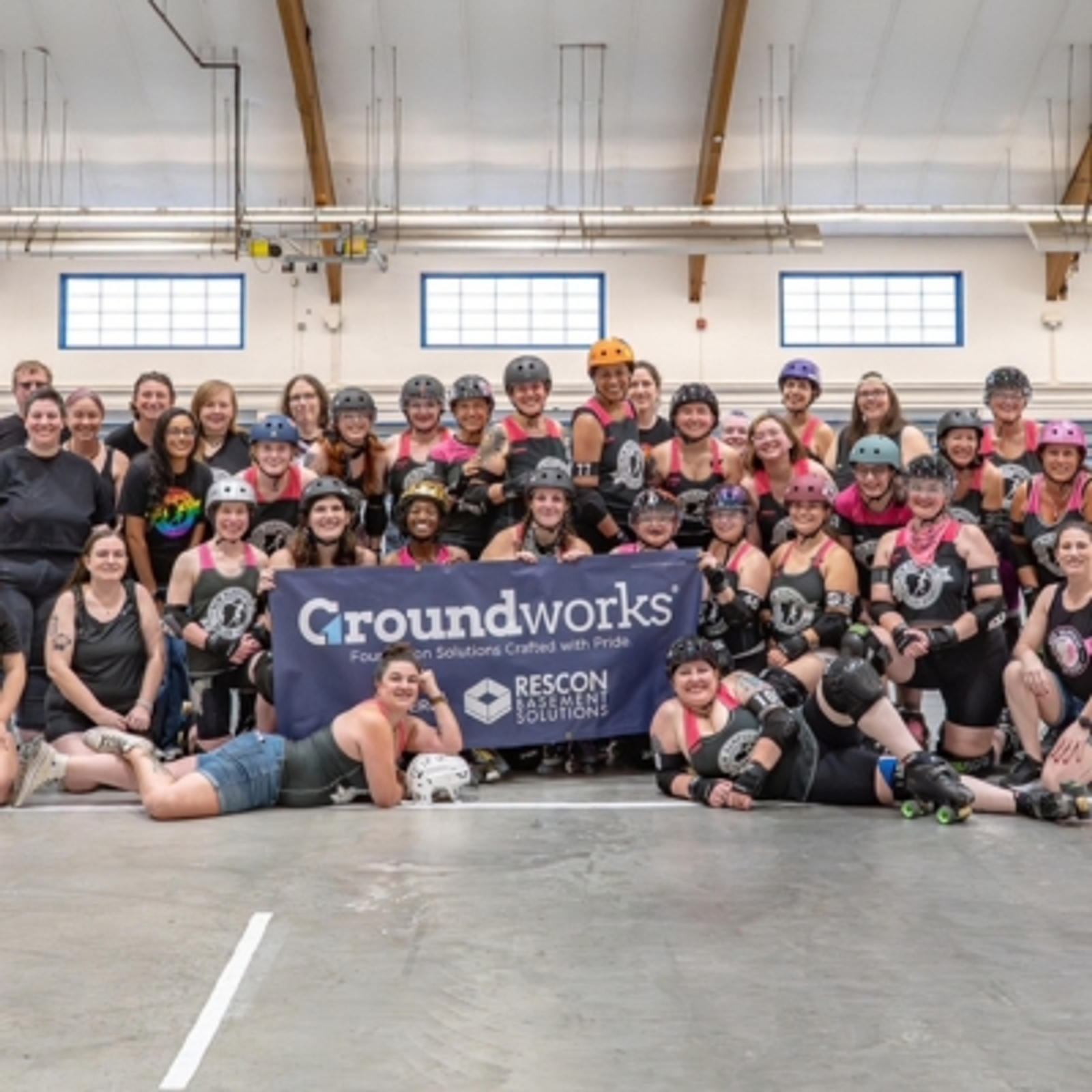 A large roller derby team poses indoors, smiling together behind a "Groundworks" sponsor banner.