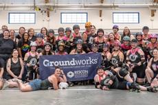 A large roller derby team poses indoors, smiling together behind a "Groundworks" sponsor banner.