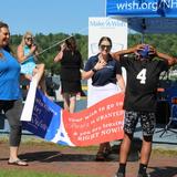 A boy in a black "4" jersey reacts with joy as adults reveal a "Make-A-Wish Paris" banner outdoors.