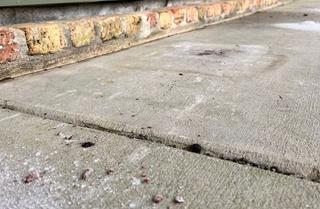 Uneven concrete pavement with a raised edge near a brick wall, showing some debris and dirt on the surface.