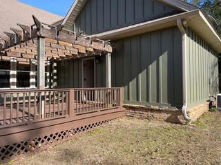 Green house with a wooden pergola and porch, brick foundation, and gutter downspout in sunlight.