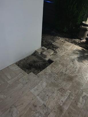 A tiled patio with a section of tiles removed and stacked, exposing dirt underneath near a white wall.