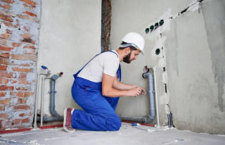 A plumber in blue overalls and a hard hat kneels, working on pipes in a partially finished room.