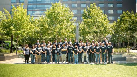 A large group of people pose for a photo outside on a sunny day, with trees and a modern building behind them.