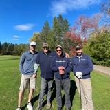 Four men stand together on a sunny golf course, smiling, with trees and autumn colors in the background.