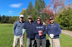 Four men stand together on a sunny golf course, smiling, with trees and autumn colors in the background.