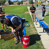 Man with a blue vest uses an air pump next to a red bucket on a grassy field; people stand nearby.
