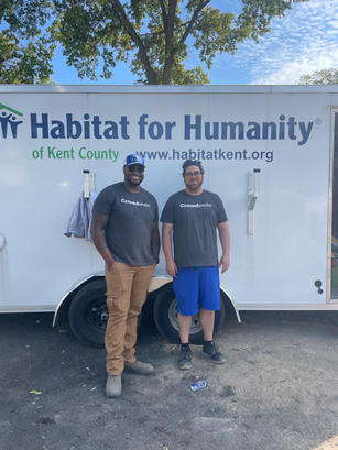 Two people smiling in front of a Habitat for Humanity of Kent County trailer on a sunny day.