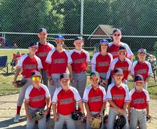 Youth baseball team in red uniforms poses together outdoors in front of a chain-link fence on a sunny day.