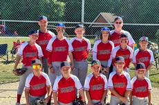 Youth baseball team in red uniforms poses together outdoors in front of a chain-link fence on a sunny day.