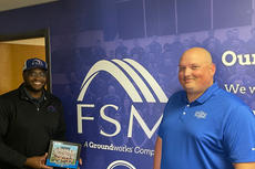 Two men standing in front of a wall with "FSM" logo, one holding a framed collage with a group photo.