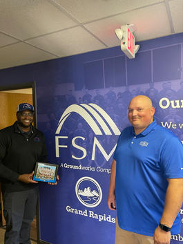 Two men standing in front of a wall with "FSM" logo, one holding a framed collage with a group photo.