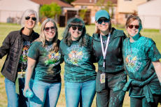 Five smiling women with sunglasses stand together outdoors, covered in blue powder, wearing matching event T-shirts.