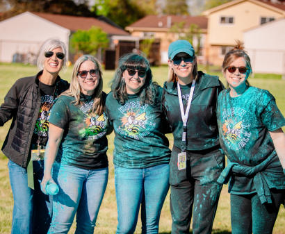 Five smiling women with sunglasses stand together outdoors, covered in blue powder, wearing matching event T-shirts.
