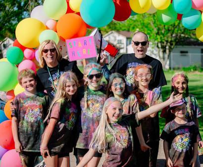 A group of kids and two adults pose under colorful balloons at a fun outdoor event, smiling and cheerful.