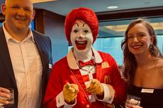 Three people smiling at an event, with Ronald McDonald standing between a man and a woman, holding drinks.