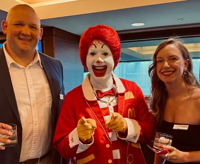 Three people smiling at an event, with Ronald McDonald standing between a man and a woman, holding drinks.