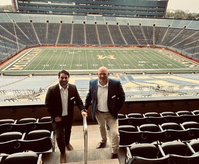 Two men in suits stand in a stadium suite overlooking an empty Michigan football field.