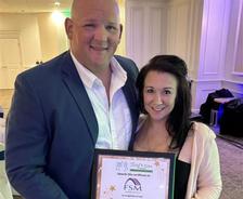 Two people smiling and holding a framed certificate while posing together in a formal indoor setting.