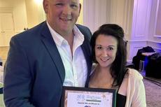 Two people smiling and holding a framed certificate while posing together in a formal indoor setting.