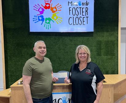 Two people standing in front of a "Macomb Foster Closet" sign, holding a check together.