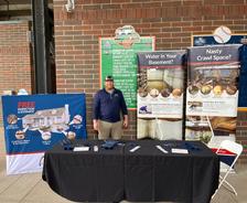 Man stands behind a table with flyers at a home inspection booth with banners and a brick wall in the background.