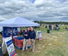 Two men smile and pose by a promotional tent at an outdoor event with people and booths in the background.