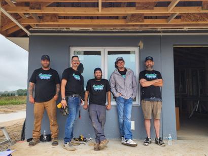 Five smiling men stand in front of a house under construction, wearing matching shirts and caps.