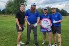 Four men standing on a golf course, one holding a circular FSM sponsor sign, all posing and smiling.