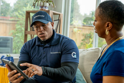 A man in a Groundworks uniform shows a woman something on a tablet while sitting together indoors.